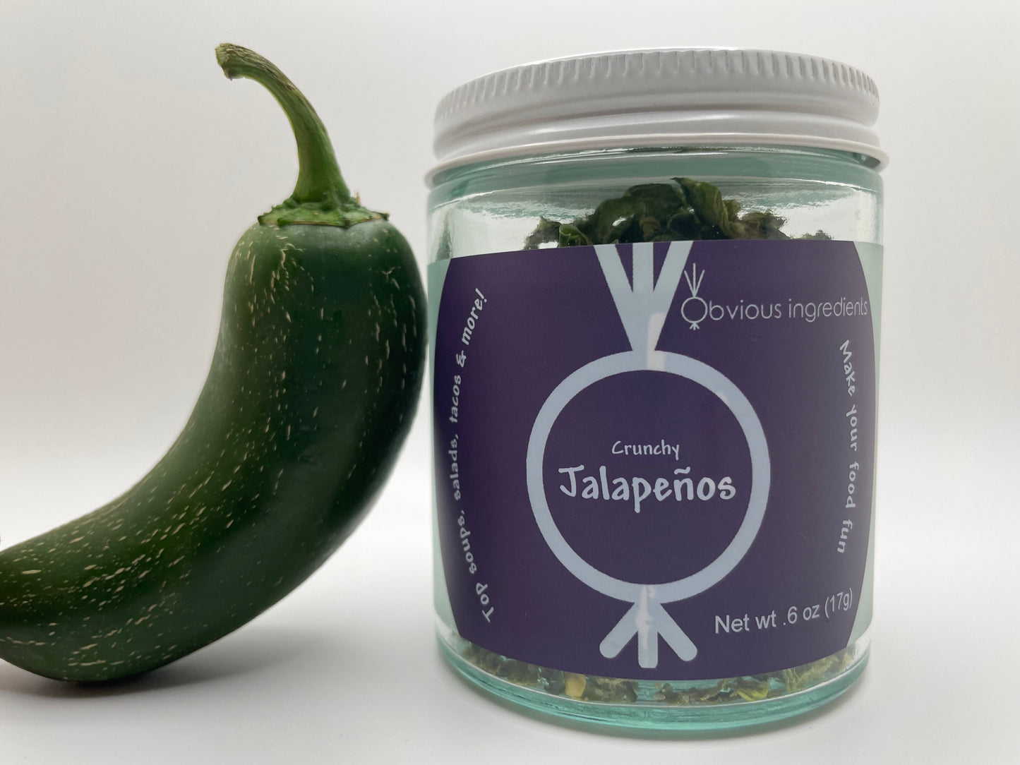 Clear jar of dried jalapeños with purple label and fresh jalapeño leaning against it