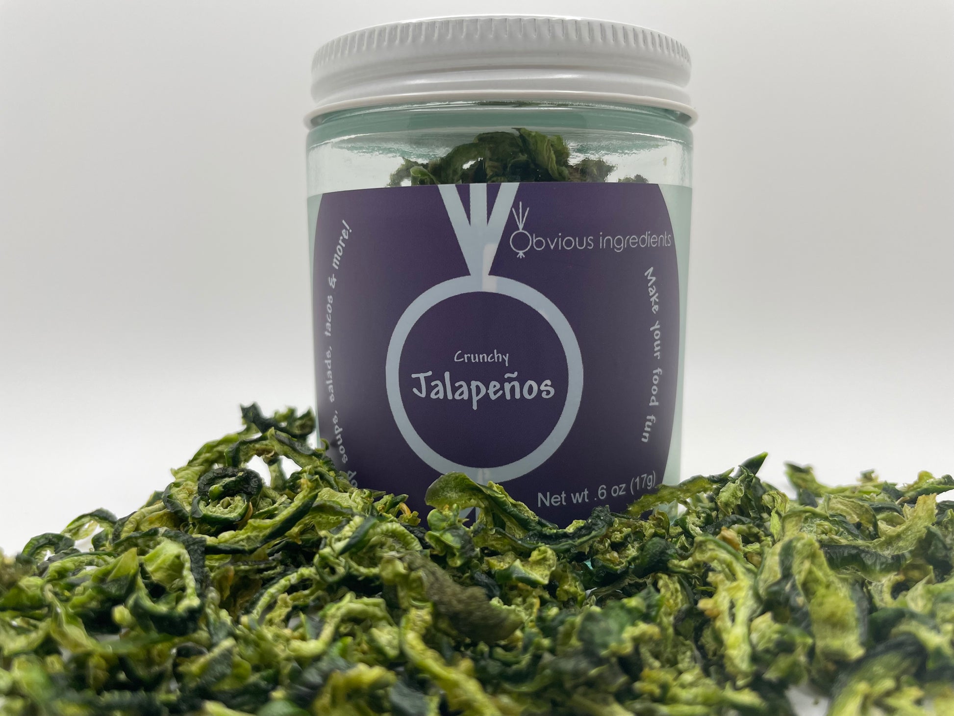 Clear jar of dried jalapenos with purple label surrounded by chopped dried jalapenos with white background